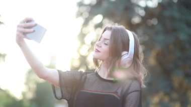 Güzel bir bayan model şehirde duruyor ve cep telefonuyla müzik dinliyor. Modern kulaklıklar kullanarak. Screen 'e yazıp popüler şarkıları seçmek. Çekici görünüş ve şık kıyafet..