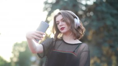 Güzel bir bayan model şehirde duruyor ve cep telefonuyla müzik dinliyor. Modern kulaklıklar kullanarak. Screen 'e yazıp popüler şarkıları seçmek. Çekici görünüş ve şık kıyafet..