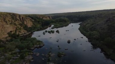 Bir dağ nehrinin havadan çekilmiş görüntüsü, bir dağın üzerinden geriye doğru uçuyor, gün batımında yeşil nehir kıyısından geçiyor, akıntılı küçük kayalar, su yüzeyinde güneş ışığı