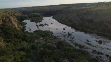 Bir dağ nehrinin havadan çekilmiş görüntüsü, bir dağın üzerinden geriye doğru uçuyor, gün batımında yeşil nehir kıyısından geçiyor, akıntılı küçük kayalar, su yüzeyinde güneş ışığı
