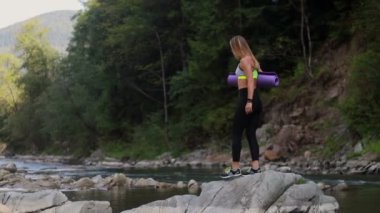 Ellerinde spor minderleriyle sabahları spor egzersizleri yapmak ve yoga yapmak için dağ nehrinin kenarında poz veren lasinas giymiş genç bir kadın.