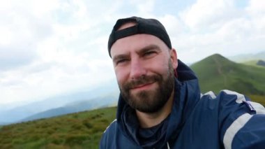 Turizm, dağlar, yaşam tarzı, doğa, insanlar, selfie konsepti - Genç Gezgin yazın, gün batımında, arka plandaki dağlarda selfie çekiyor. Sakallı turist gülümsüyor. Ağır çekim, teknoloji