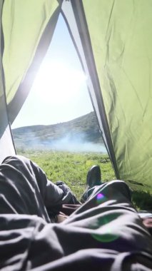 Adam çadırın içinde dinleniyor, bacak bacak üstüne atıyor, ayaklarına bakıyor, arka planda güzel örtülü bir dağ var.