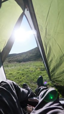 Adam çadırın içinde dinleniyor, bacak bacak üstüne atıyor, ayaklarına bakıyor, arka planda güzel örtülü bir dağ var.