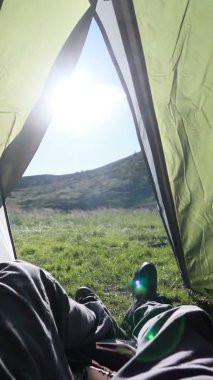 Adam çadırın içinde dinleniyor, bacak bacak üstüne atıyor, ayaklarına bakıyor, arka planda güzel örtülü bir dağ var.