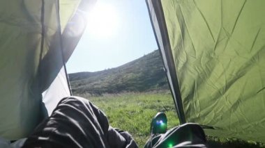 Adam çadırın içinde dinleniyor, bacak bacak üstüne atıyor, ayaklarına bakıyor, arka planda güzel örtülü bir dağ var.