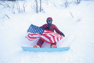 kış, eğlence, spor ve insan konsepti - snowboardcu yamacın kenarındaki dağlarda oturur ve uzaklığa bakar. Amerikan bayrağı taşıyan snowboardcu