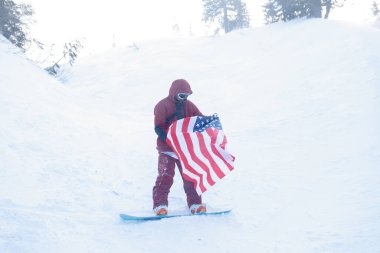 kış, eğlence, spor ve insan konsepti - snowboardcu yamacın kenarındaki dağlarda oturur ve uzaklığa bakar. Amerikan bayrağı taşıyan snowboardcu