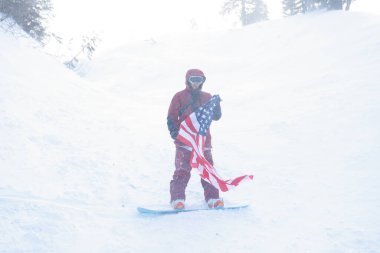 kış, eğlence, spor ve insan konsepti - snowboardcu yamacın kenarındaki dağlarda oturur ve uzaklığa bakar. Amerikan bayrağı taşıyan snowboardcu