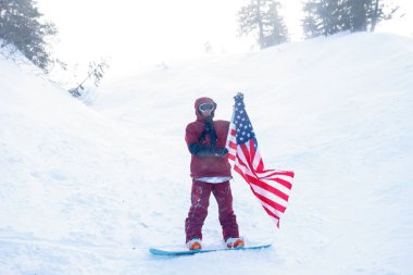 kış, eğlence, spor ve insan konsepti - snowboardcu yamacın kenarındaki dağlarda oturur ve uzaklığa bakar. Amerikan bayrağı taşıyan snowboardcu