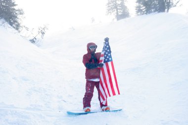 kış, eğlence, spor ve insan konsepti - snowboardcu yamacın kenarındaki dağlarda oturur ve uzaklığa bakar. Amerikan bayrağı taşıyan snowboardcu