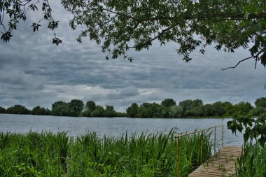 Küçük bir balıkçı köprüsü, bir nehrin kıyısındaki küçük bir rıhtım ya da uzun otların arasındaki çalılıklarda, karşı kıyıların arka planında, güzel desenli bulutları olan alçak gökyüzü.