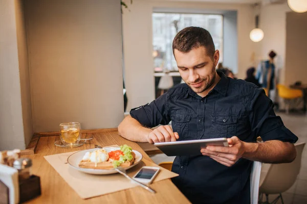 테이블에 앉아서 일 하는 실업가의 그림. 그는 태블릿을 사용 하는. 맛 있는 음식과 음료는 테이블에 접시가 있다. 또한 플레이트 근처 거짓말 전화는. — 스톡 사진