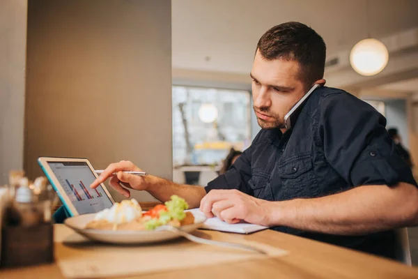 Una foto del hombre de negocios cenando en la cafetería. Está trabajando ahora mismo. Guy está hablando por teléfono y apuntando en la tableta con gráficos en su pantalla. Hay un plato con comida en la mesa . — Foto de Stock