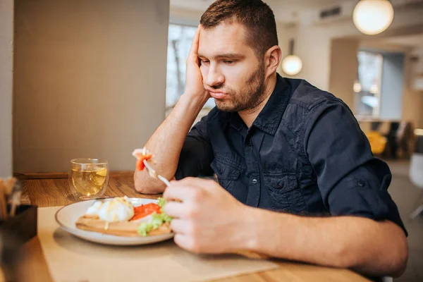 Bored and sad man is sitting at table and cafe. He is holding a piece of vegetable on fork. Man is looking at it and breathing out. — Stock Photo, Image