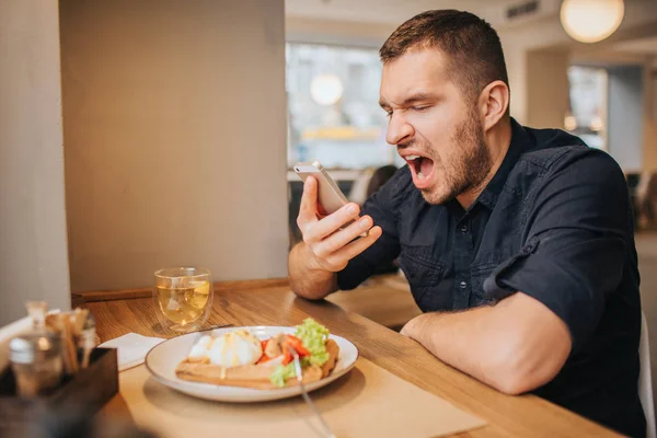 Šílený muž sedí u stolu a křičí do telefonu. Je rozzlobený a nešťastná. Chlap nemůže přestat ječet. Nejí jídlo. — Stock fotografie
