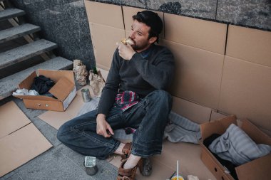 Koyu renk saçlı ve kirli adam karton üzerinde oturan ve sandviç yeme. Onun poz rahat. Adam arıyor. Onu çevreleyen bir sürü olduğunu