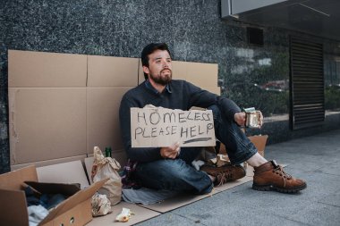 Evsiz adam karton üzerinde oturan ve ve evsizlere yardım lütfen yazan bir tabela holding. Para yalvarıyor. Adam umutsuzluk içinde ve çok yardıma ihtiyacı var.