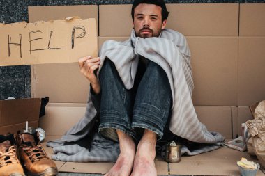 Adam yere oturuyor ve buz gibi. Onun omuzlarında küçük bir battaniye var. Adam yalındır görünüyor. Halen yardım diyor karton parçası tutuyor. Bir yardıma ihtiyacı.