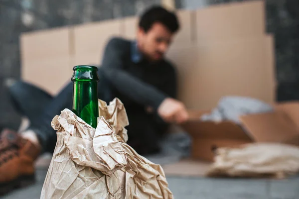 Camera is concentrated on green bottle standing in front of guy whos sitting on the cardboard and reaching the box. Body of bottle is covered with paper. — Stock Photo, Image