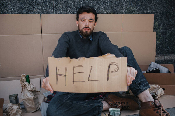 Portrait of homeless guy sitting on cardboard and holding a help cardboard in hands. He is looking straight at camera. There are lots of stuff and paper surrounding man.