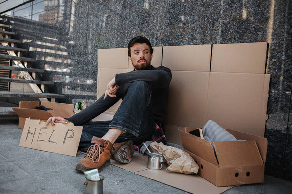 Sad and tired dark-haired man is sitting on the cardboard and holding another cardboard with the word help writing on it. He is looking aside. He is homeless.