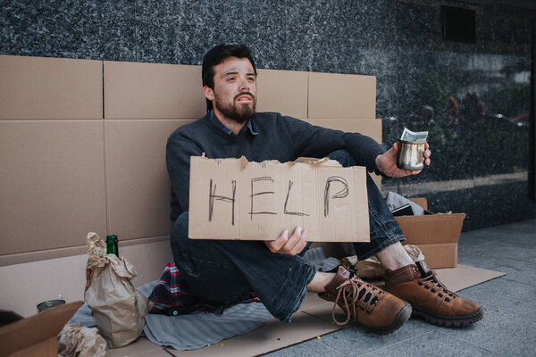 Dark-haired man is sitting on the cardboard and holding a sign that says help. He is holding in left hand a metal cup with one dollar in it. Guy has lots of stuff lying besides him.