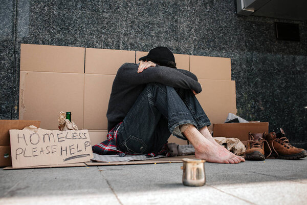 A picture of hopeless man sitting on the cardboard and hiding his face. He is covered it with hands and looking down. Man is sitting without shoes on. He is a beggar. There is metal cup for charity.