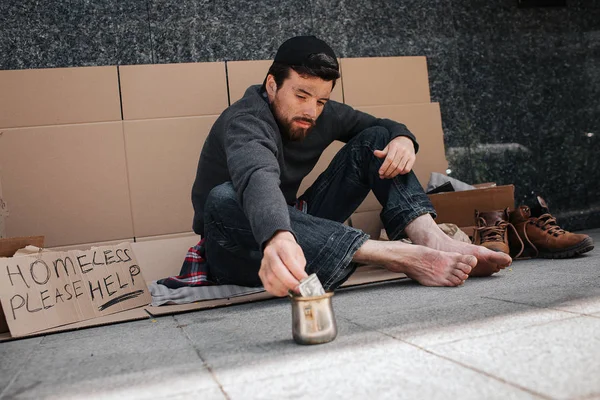 Hemlös man sitter på den kartong utanför och nå metall koppen med handen. Han når en dollar. Det finns en skylt säger hemlösa snälla hjälp. Killen vill ta pengar. — Stockfoto