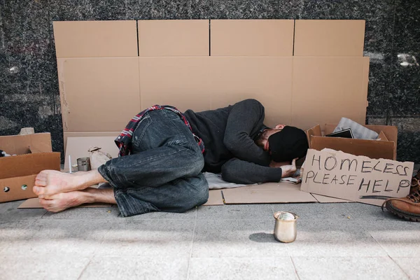 Hjälplös och försvarslösa mannen är liggande på kartong på betonggolv och sover. Han täcker ansiktet med händerna dölja det från solen. Det finns ett tecken förutom honom säger hemlösa snälla hjälp. — Stockfoto