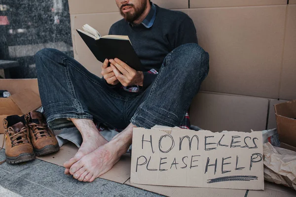 Smart hemlös man sitter på kartong och läsa en bok. Han är mycket koncentrerad på det. Det finns en suck som säger hemlösa snälla hjälp. — Stockfoto