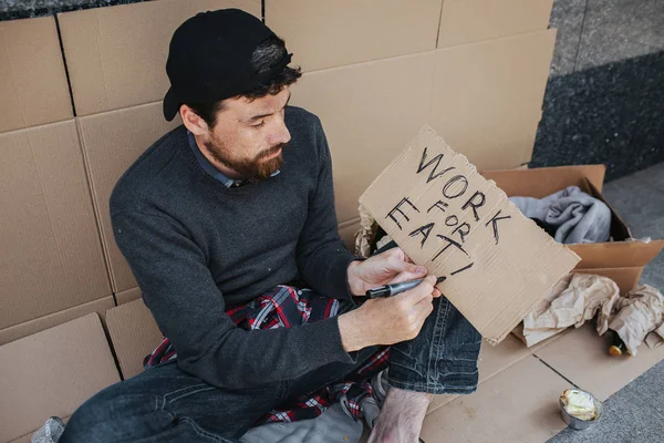 Arbetslösa man sitter på marken och skriver ner ord arbete för äter på papper. Kille söker ett jobb. Han hoppas att någon kommer att hjälpa honom. — Stockfoto