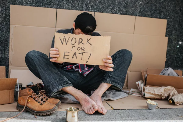 Arbetslösa kille sitter på betong marken och som täcker ansiktet med tecken arbete för eat. Han söker ett jobb. Killen är mycket dålig. Han hoppas att någon kommer att hjälpa honom. — Stockfoto