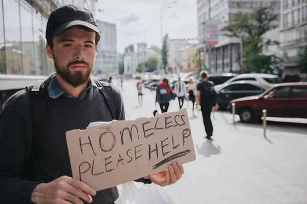 Hombre sin hogar está parado en la calle y mostrando el cartel que dice sin hogar por favor ayuda. El tipo está esperando ayuda de alguien. Parece serio y cansado. . —  Fotos de Stock