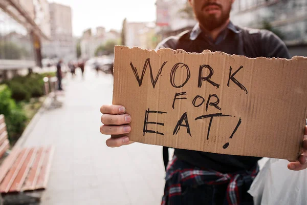 En bild av hemlös man står utanför och hålla en kartong som säger arbete för eat. Kille inte har ett jobb och letar efter den. — Stockfoto