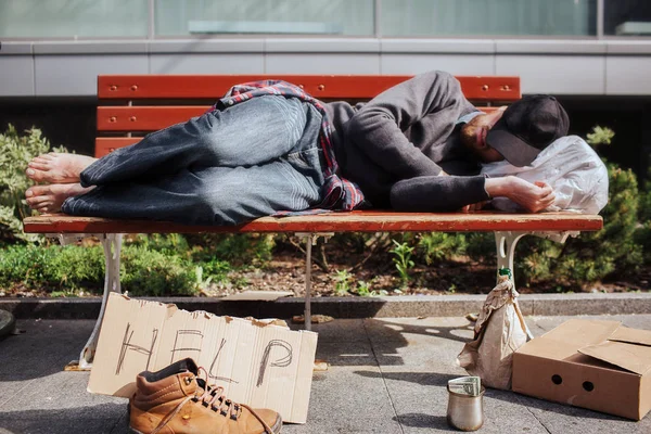 Hemlös man är liggande på bänken och sover. Hee är trött och utmattad. Det finns en väska med grejer under hans huvud. Det finns en hjälp-kartong och en metall kopp med pengar på marken. — Stockfoto
