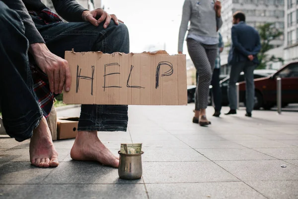En bild av mans ben utan skor står på betong marken. Det finns en metall kopp med pengar på den. Killen håller en kartong som säger hjälp. — Stockfoto