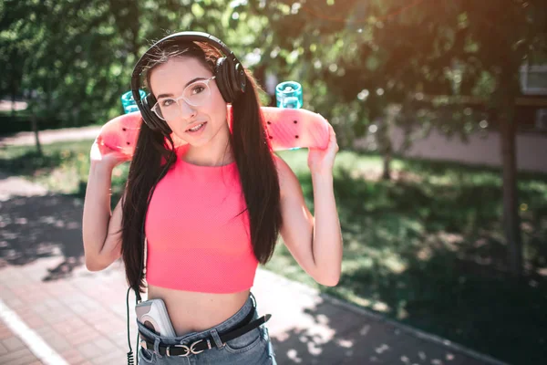 Mulher atraente em camisa rosa está posando. Ela está olhando diretamente para a câmera e segurando patins atrás da cabeça. Também menina está ouvindo música através de fones de ouvido usando o jogador para que . — Fotografia de Stock