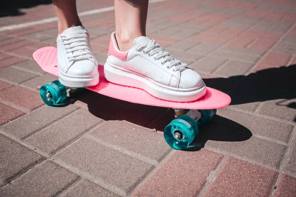 Primer plano de las niñas piernas y pies lleva zapatillas blancas. La chica está parada en el patín rosa con ambos pies. Hay un día soleado afuera. . —  Fotos de Stock