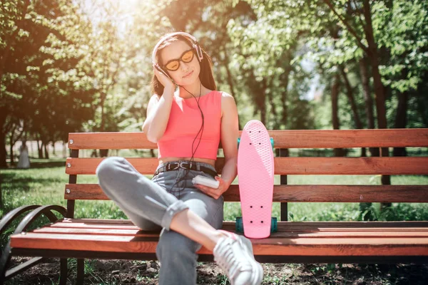 Fille heureuse profite de l'instant. Elle écoute de la musique à travers des écouteurs et profite du moment. Elle a mis la jambe droite sur la gauche. Il y a un patin à roulettes debout sur le banc . — Photo