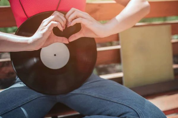 Een foto van zwarte vynil leunend naar meisjes lichaam. Ze is zittend op de Bank en haar benen oversteken. Meisje is shwoing de liefde ondertekenen met haar vingers voor vinyl schijf. — Stockfoto