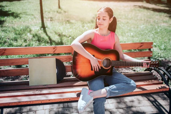 A rapariga simpática está sentada no banco e a tocar guitarra. Ela está olhando para baixo para o vynil e sua capa que está no banco também. Ela está desfrutando o momento . — Fotografia de Stock