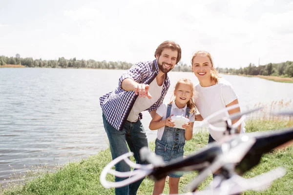 Magnifique famille est debout ensemble et regardant drone qui est sur la photo. Petite fille a panneau de commande et souriant. Un jeune homme pointe du doigt un drone . — Photo