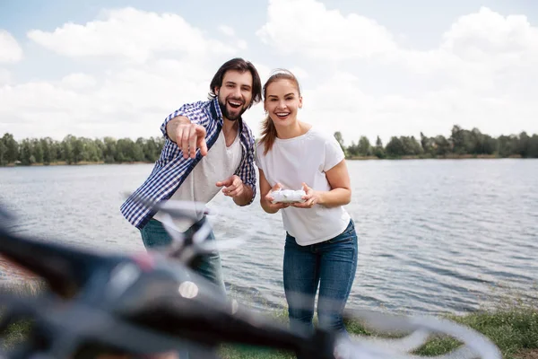 Nice and happy couple is standing together and launching drone. It is contlolled by control panel. Man is pointing on drone. They are excited. — Stock Photo, Image
