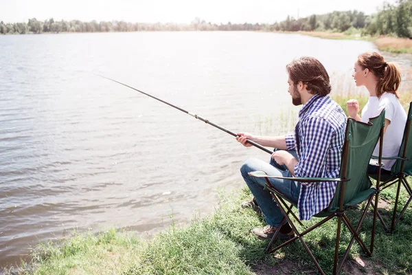 Une photo d'un homme et d'une femme adultes assis au bord de la rivière par cette belle et belle journée ensoleillée. Il essaie d'attraper du poisson. Tous les deux regardent vers l'eau . — Photo