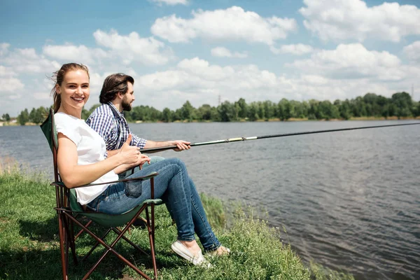 Ευτυχισμένη γυναίκα κάθεται στην μαλακή καρέκλα και ψάχνει για την κάμερα. Είναι χαμογελά. Ο σύζυγός της συνεδρίαση εκτός της και προσπαθεί να πιάσει μερικά ψάρια χρησιμοποιώντας ψάρια-rod. — Φωτογραφία Αρχείου