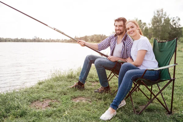 Una foto de jóvenes increíbles sentados juntos en la orilla del río y sonriendo en cámara. Chico está sosteniendo enorme y larga caña de pescado mientras su esposa se inclina la cabeza sobre su hombro . — Foto de Stock