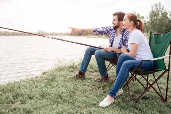 Dos personas están sentadas en la orilla del río. El tipo sostiene el borde de la caña de pescar y señala el agua mientras la chica mira en la misma dirección que el chico. Son muy tranquilos y pacíficos. . — Foto de Stock