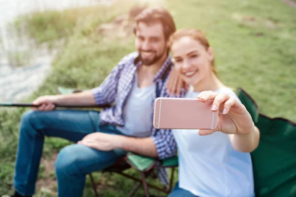 Dziewczyna jest biorąc selfie z siebie i męża. Oni są patrząc na telefon i uśmiecha się. Facet trzyma na końcu wędki na duże ryby. Ludzie siedzą w składane krzesła. — Zdjęcie stockowe