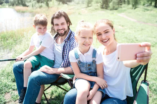 La mujer está sentada junto con su familia y tomando fotos de ella. Todos ellos están mirando a la cámara y sonriendo. Niño pequeño está sentado en su regazo papás y sosteniendo la caña de pescado . — Foto de Stock